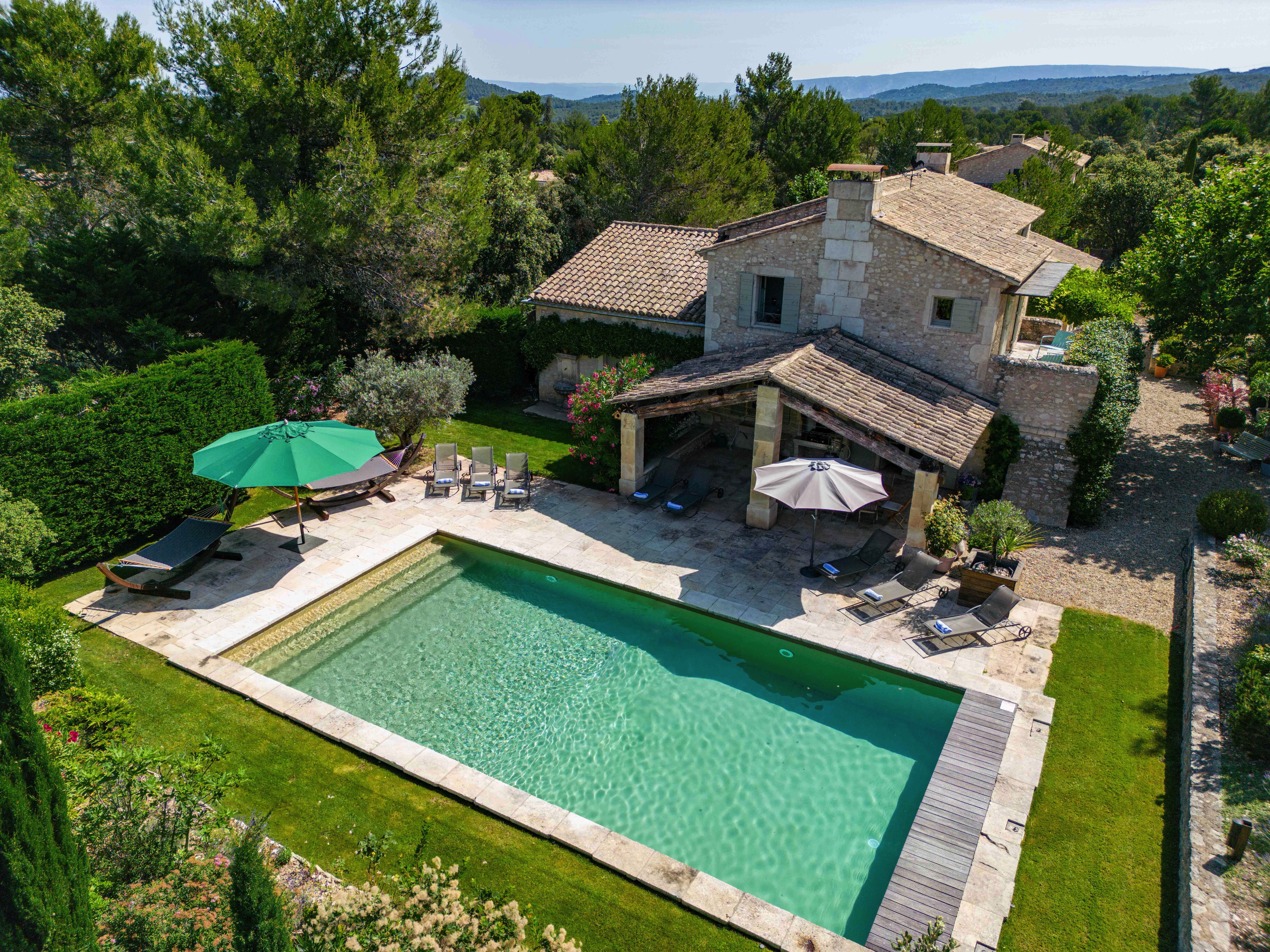 Aerial view of a luxurious house with a swimming pool, surrounded by lush greenery and equipped with patio areas and umbrellas, in a serene, sunny landscape.